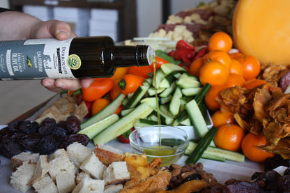 Olive oil being poured over a grazing board of vegetables, bread and fruit