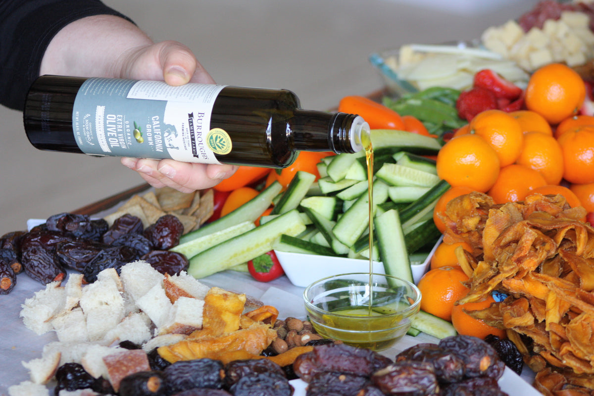 Olive oil being poured over a grazing board of vegetables, bread and fruit