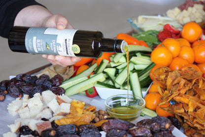 Olive oil being poured over a grazing board of vegetables, bread and fruit