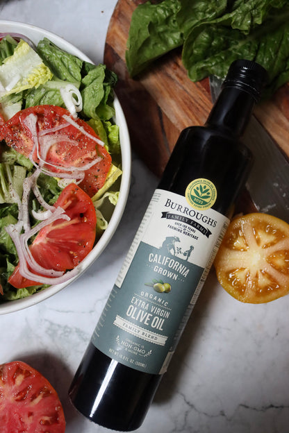 Bottle of Family Blend olive oil beside a bowl of salad and sliced tomatoes
